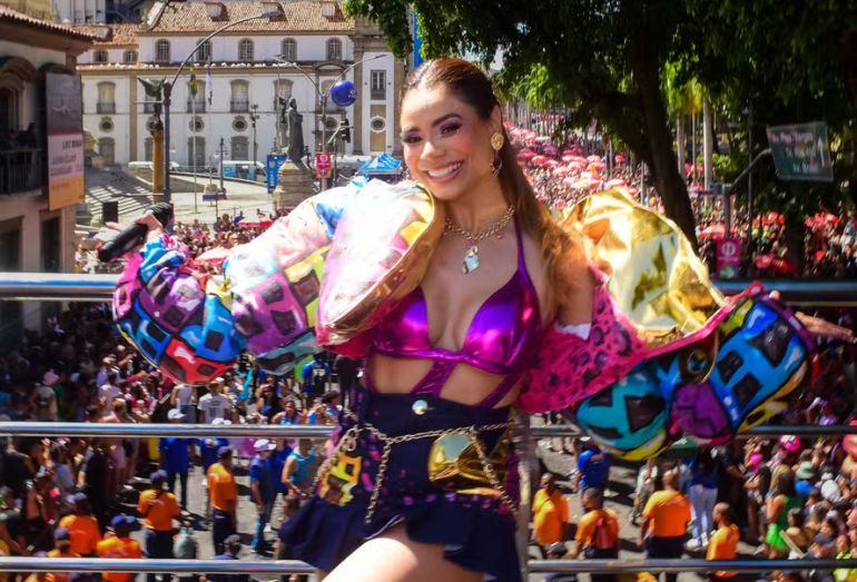 A cantora Lexa com um figurino colorido e brilhante, segurando o microfone no alto de um trio elétrico cercado por uma multidão no Centro do Rio de Janeiro.