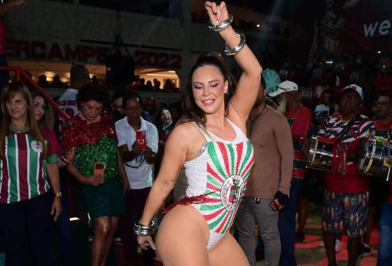 A atriz Paolla Oliveira sorrindo e sambando, vestindo um maiô cavado nas cores verde e vermelho, com sandálias de amarração verde, na quadra da escola de samba.